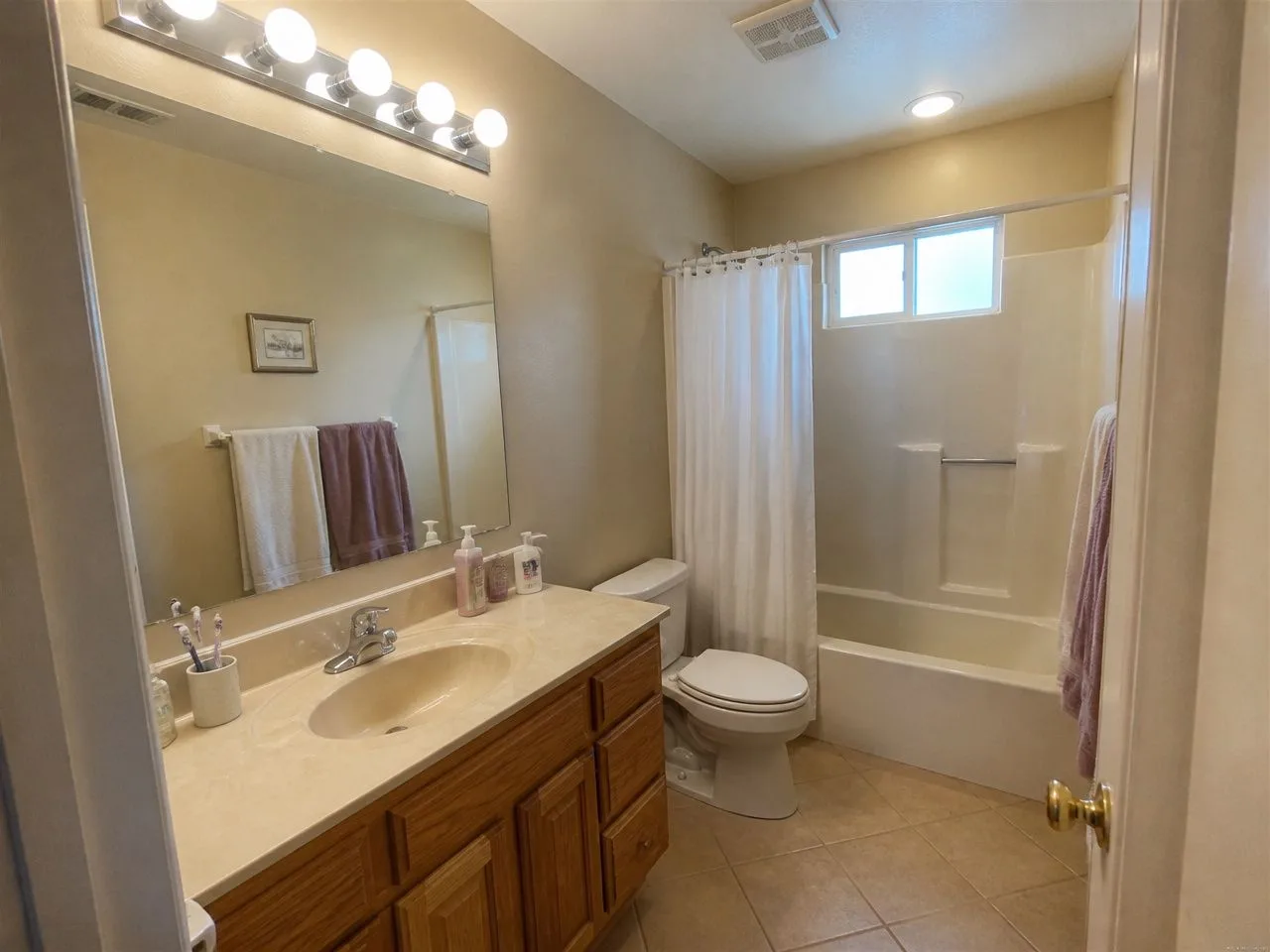 A dated 1990s hall bathroom with an oak vanity, a Hollywood light bar and an almond tub-shower, before the AI redesign
