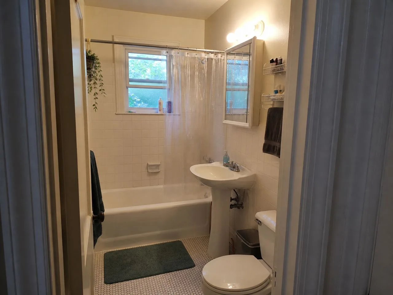 A small rental bathroom with white square tile, a tub with a plastic curtain and a pedestal sink